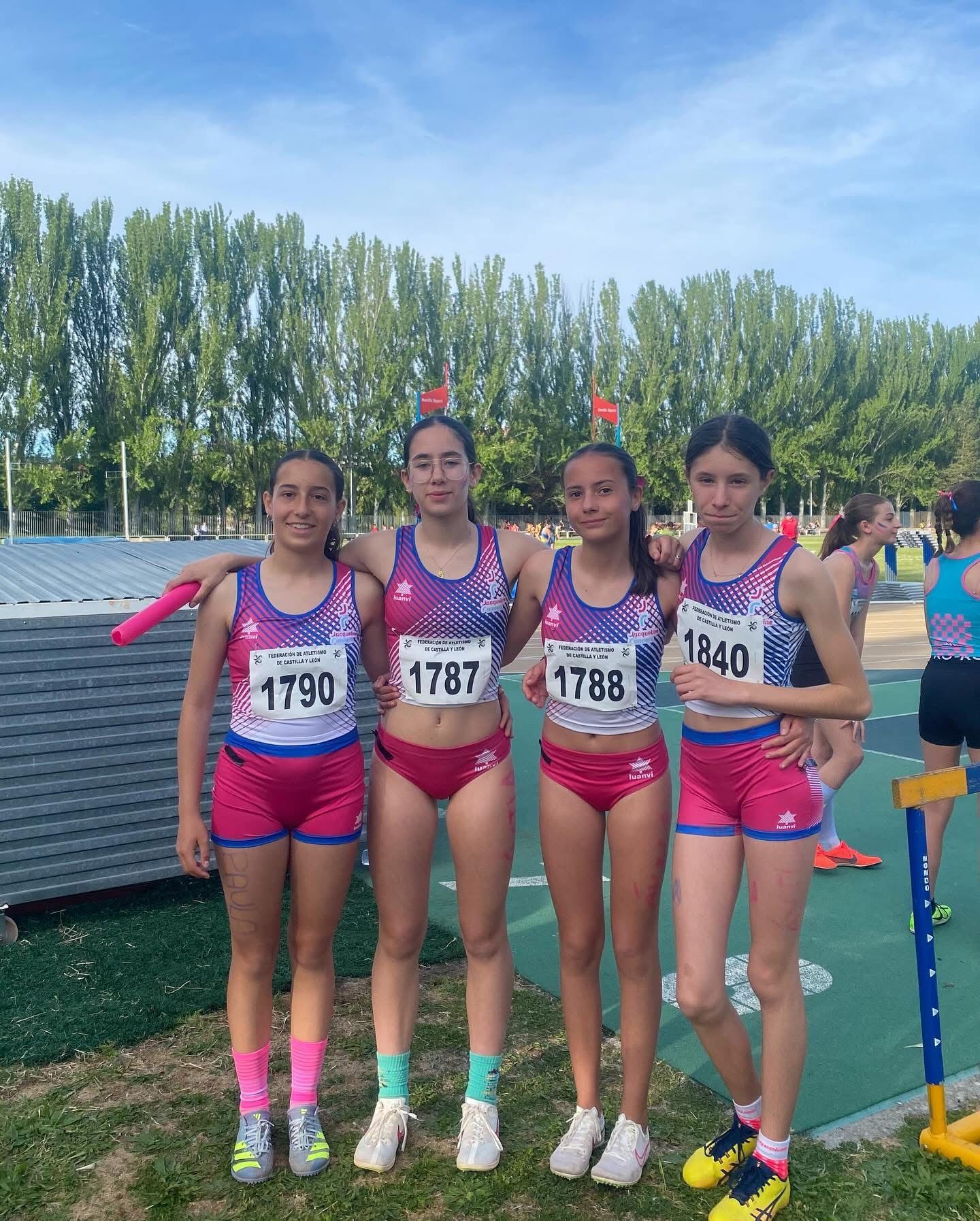 Cuatro jóvenes atletas con uniformes de atletismo posan en la pista. Lucen uniformes rosas y azules