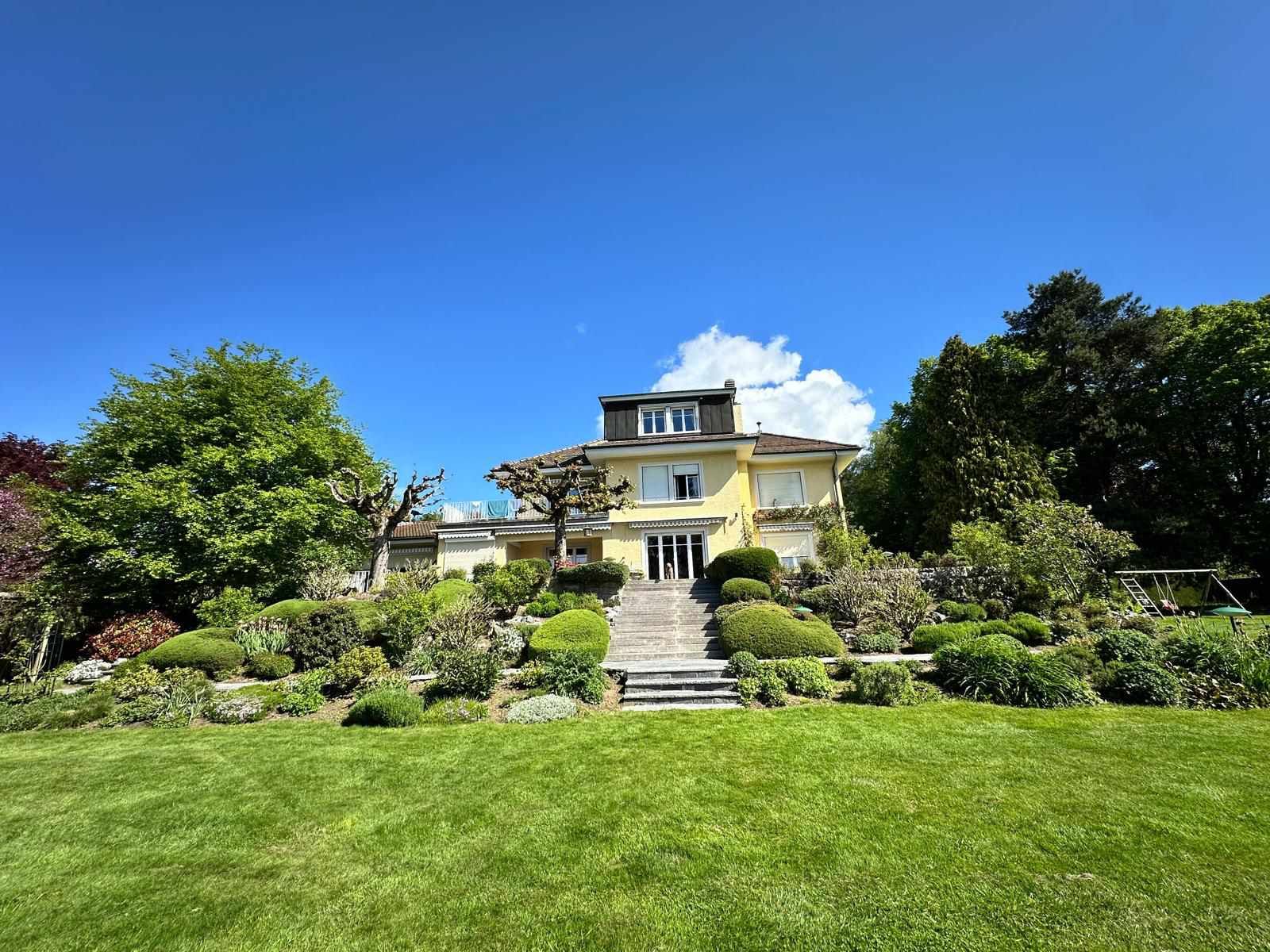 Une grande maison est située au sommet d'une colline verdoyante entourée d'arbres et de buissons.