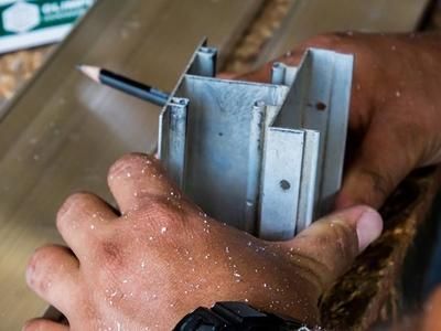 Des mains tenant une équerre métallique et un crayon pour mesurer ou marquer une pièce d’aluminium.