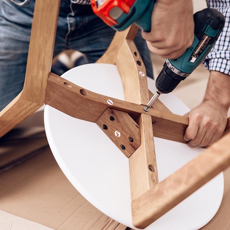 Des mains utilisant une perceuse pour assembler le cadre d’une chaise en bois avec une assise blanche.