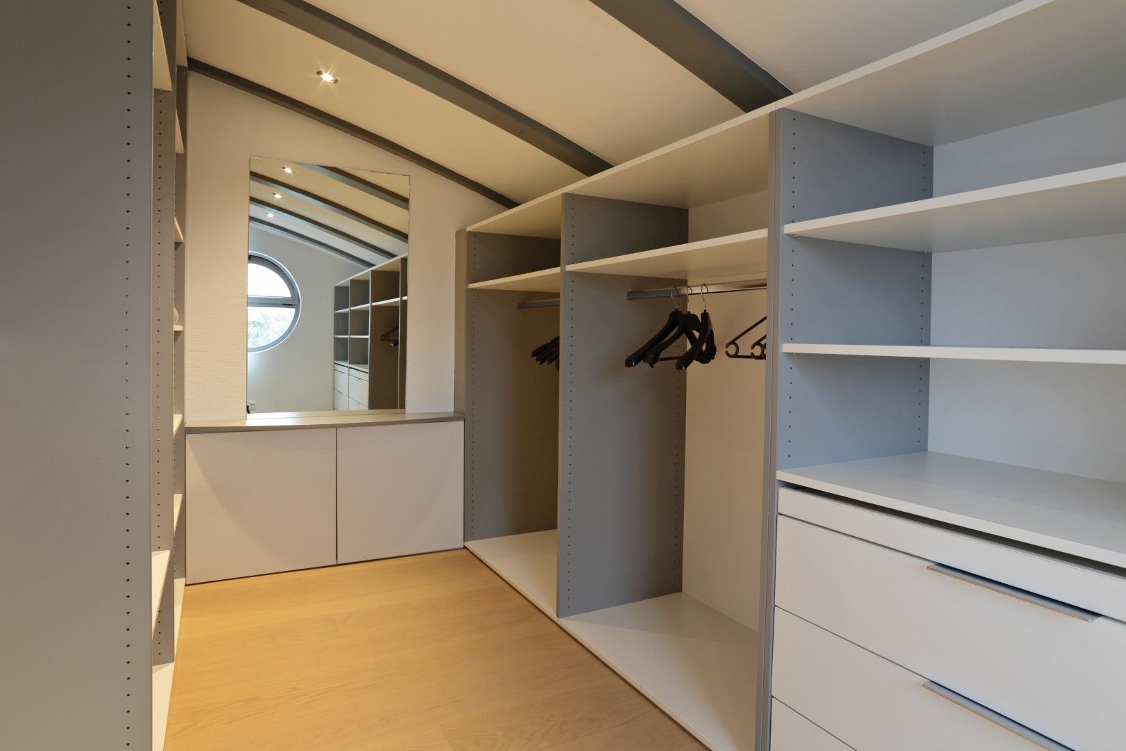 Dressing moderne avec étagères blanches, tringles à vêtements, tiroirs et mur miroir sous un plafond mansardé