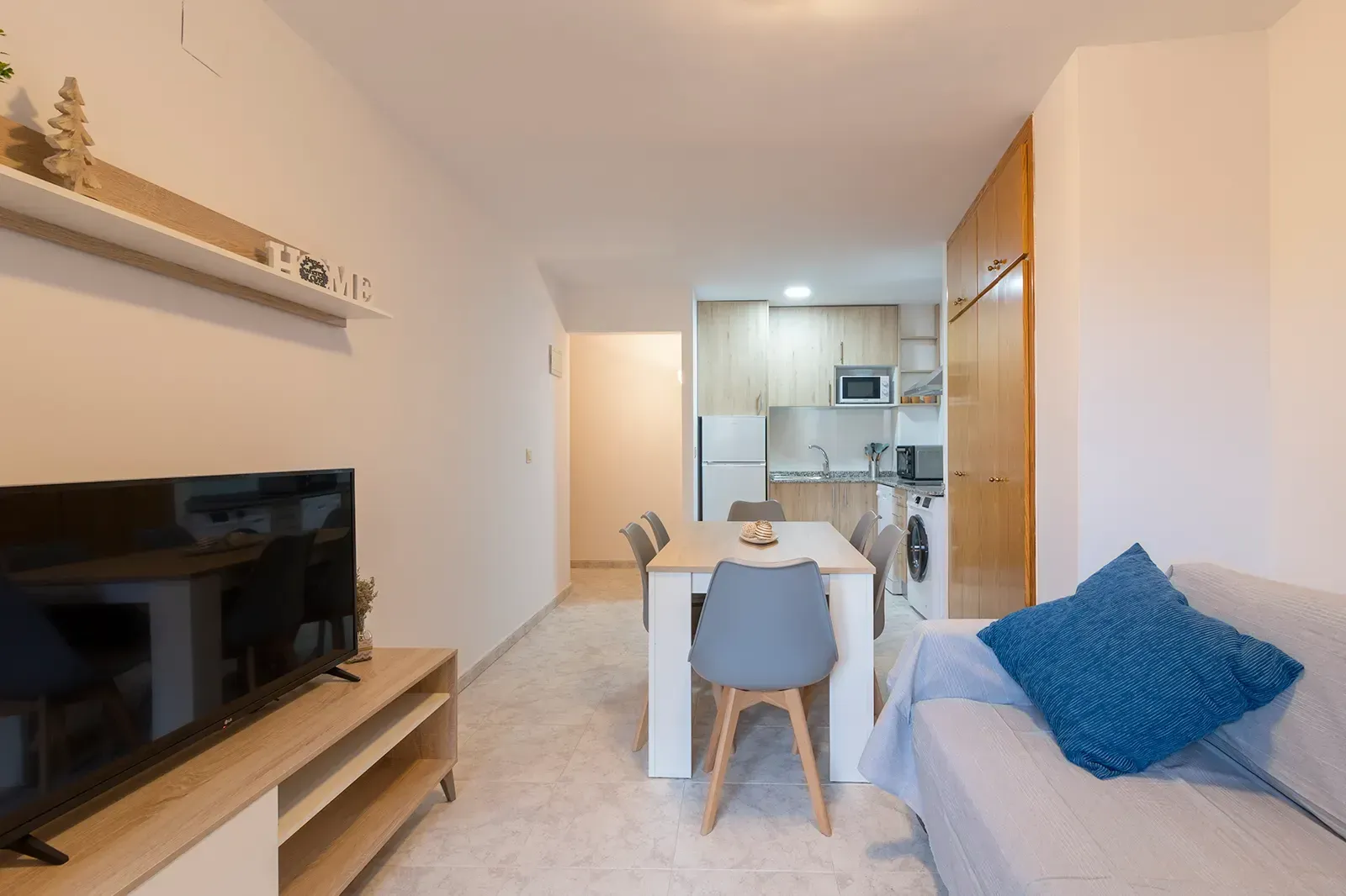 El interior de un apartamento moderno y minimalista cuenta con un televisor, un juego de comedor y un sofá con un cojín azul.