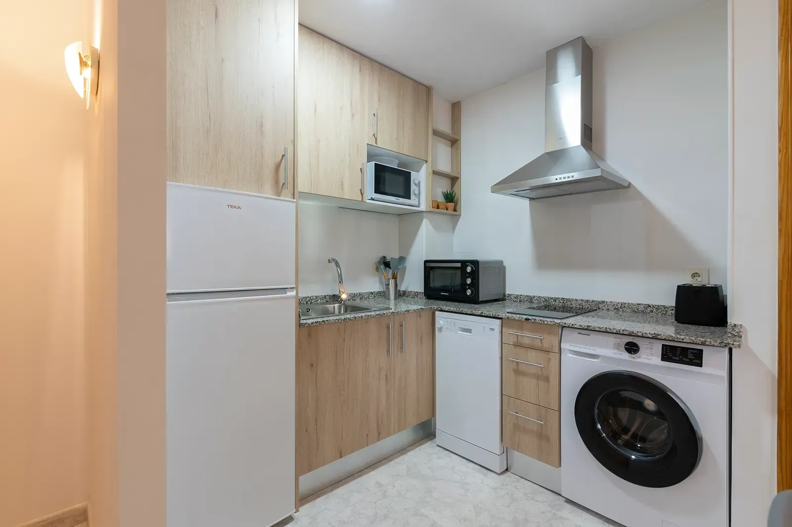 Una cocina compacta y moderna con armarios de madera clara, electrodomésticos blancos y una encimera con diseño de mármol.