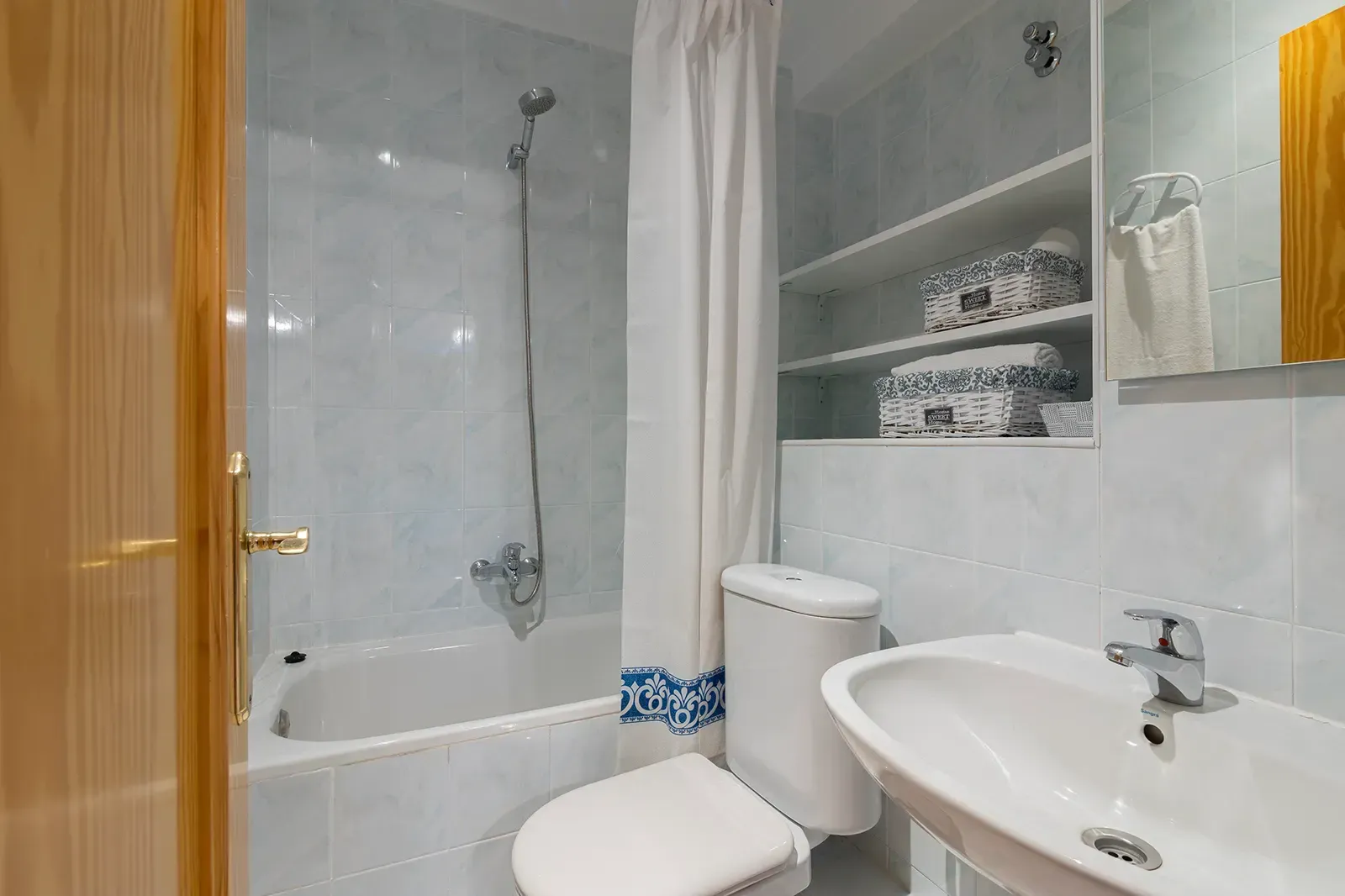 Un pequeño baño con inodoro blanco, lavabo, bañera y pilas de toallas con estampado azul en estantes abiertos en la pared.