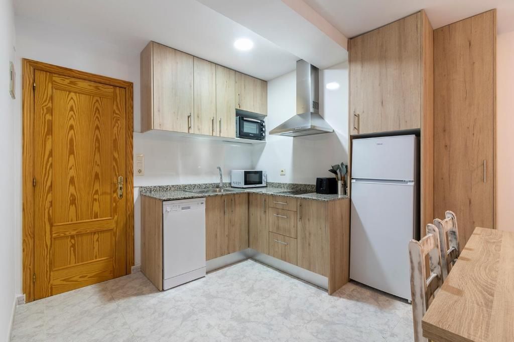 Pequeña cocina con armarios de madera clara, encimeras de granito, lavavajillas blanco y frigorífico