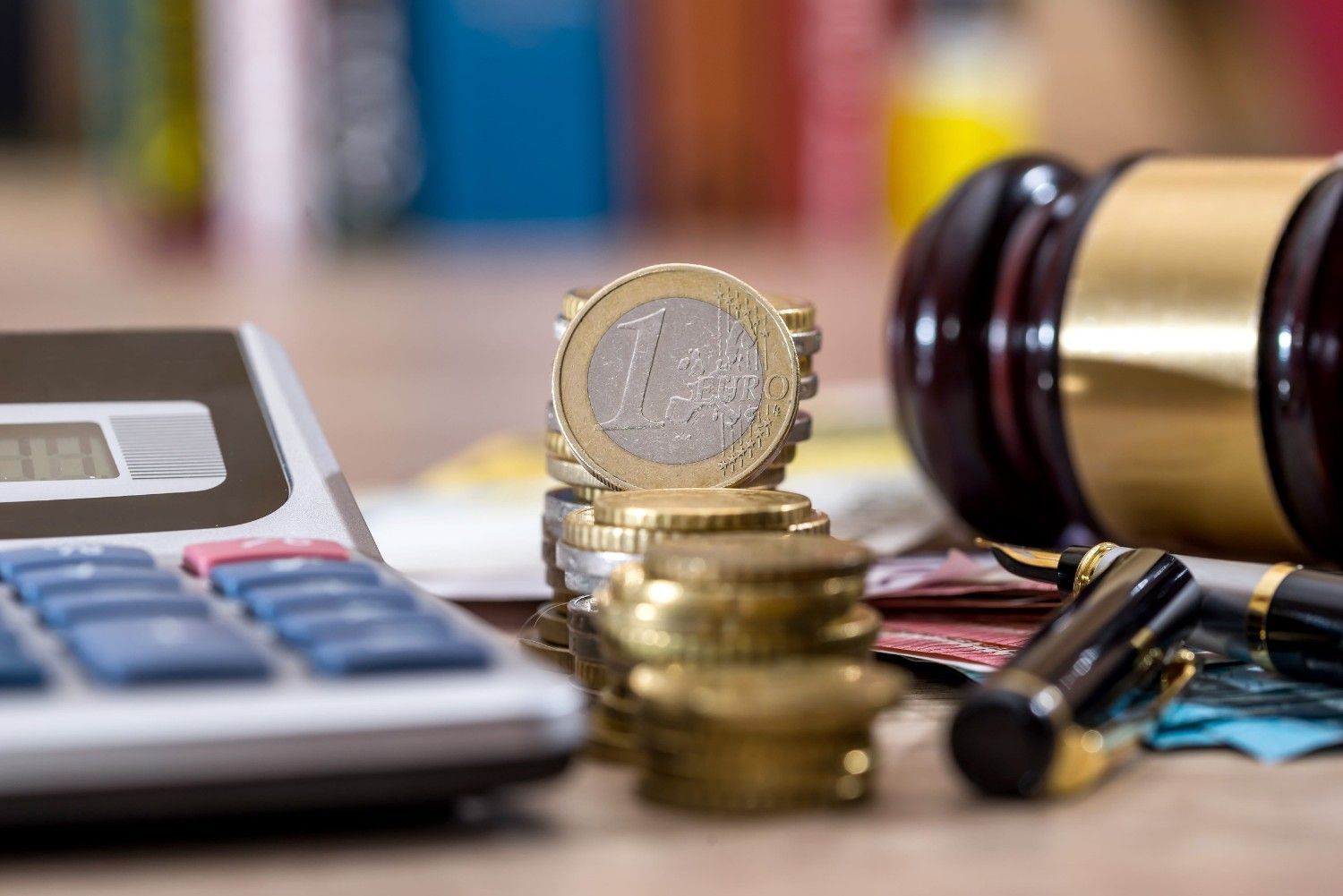 Sobre un escritorio hay una calculadora, un fajo de monedas de euro, una pluma estilográfica y un mazo de juez de madera.