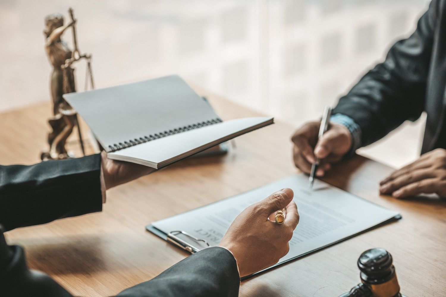 Un abogado sostiene un cuaderno abierto mientras un cliente firma un documento en un escritorio con un mazo y una estatua