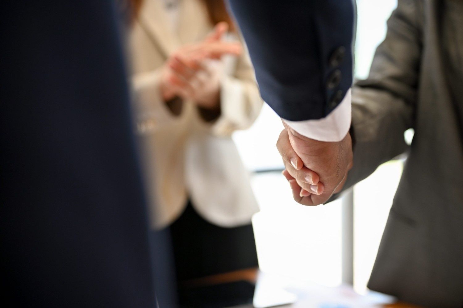Dos personas vestidas de traje de negocios se dan la mano, con una persona al fondo, en un entorno de oficina profesional.