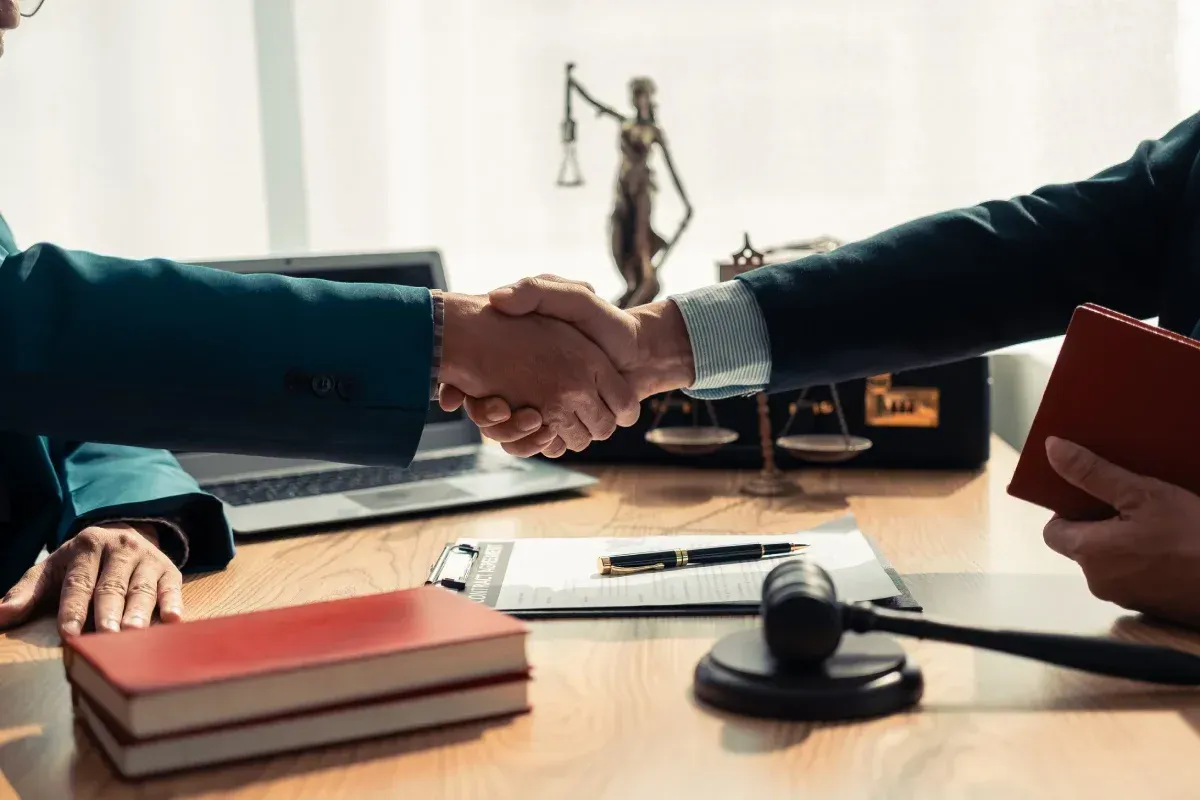 Dos personas vestidas de traje se dan la mano sobre un escritorio de madera con un mazo de juez, libros y una balanza