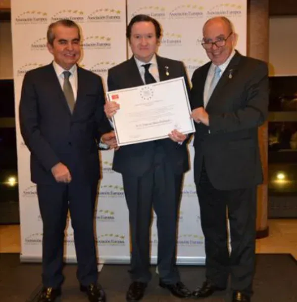 Tres hombres con traje están de pie en fila; el que está en el centro sostiene un certificado enmarcado