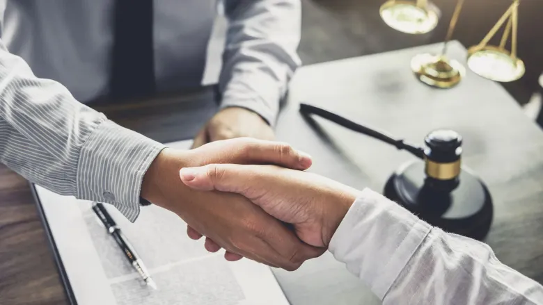 Dos personas se dan la mano al hablar de un documento legal, con el mazo del juez y la balanza de la justicia
