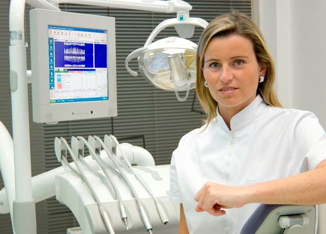 Mujer con bata blanca, apoyada en un equipo dental. Entorno profesional con pantalla digital.