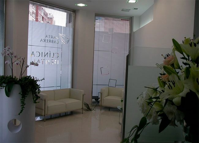 Moderna sala de espera de clínica con muebles blancos y arreglos florales cerca de las ventanas.