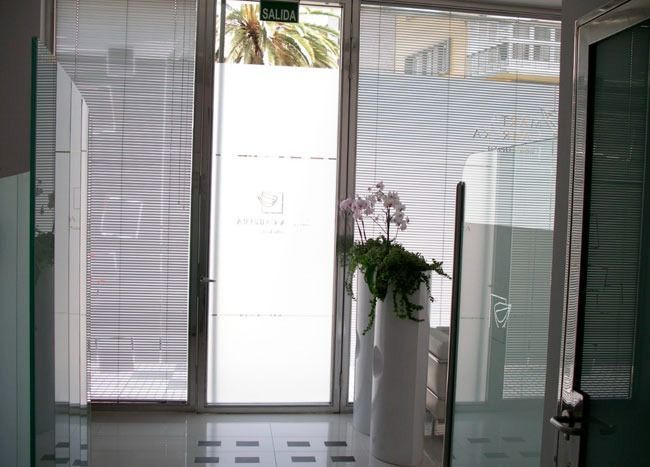 Puertas de cristal con persianas. En el interior hay un vestíbulo con plantas y letreros esmerilados.