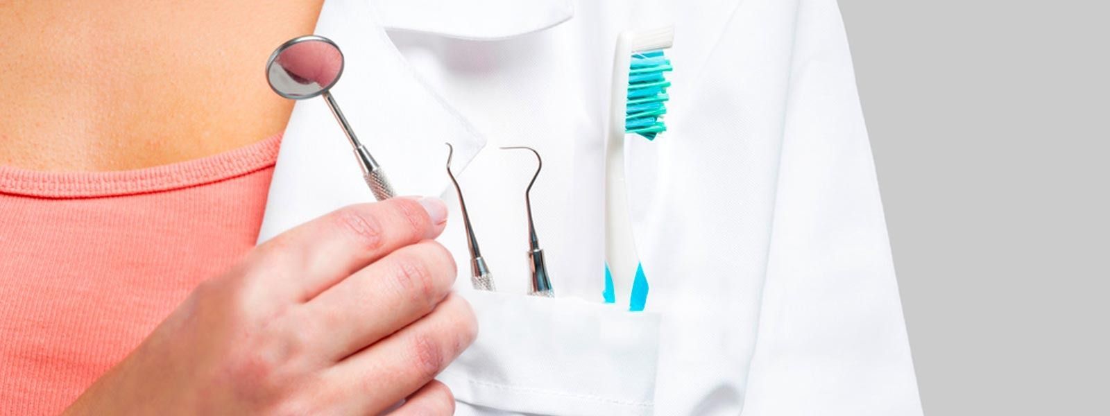Persona sosteniendo un espejo dental junto a una bata blanca con herramientas dentales y un cepillo de dientes en el bolsillo.