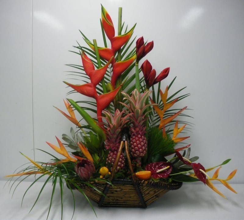 Composition florale tropicale en panier, comprenant des héliconias rouges, des petits ananas et des fleurs orange.