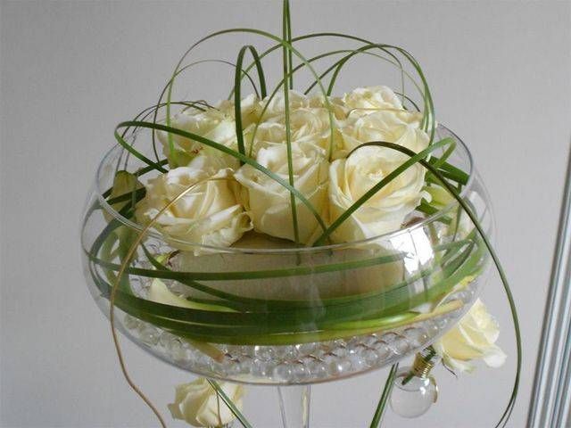 Des roses blanches dans un centre de table en coupe de verre avec des feuilles vertes ; un piédestal transparent avec des perles d'eau.