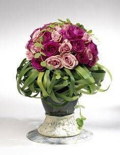 Bouquet de roses rose foncé et rose poudré sur un socle tressé vert, dans un vase sur piédestal.