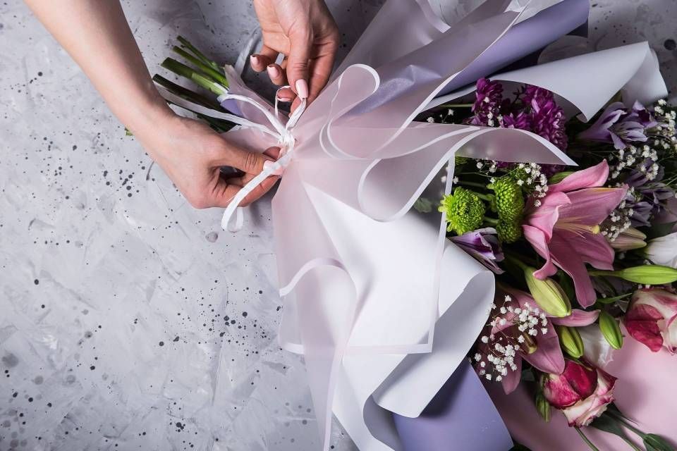 Des mains nouent un ruban blanc autour d'un bouquet de fleurs emballé, posé sur une surface grise.