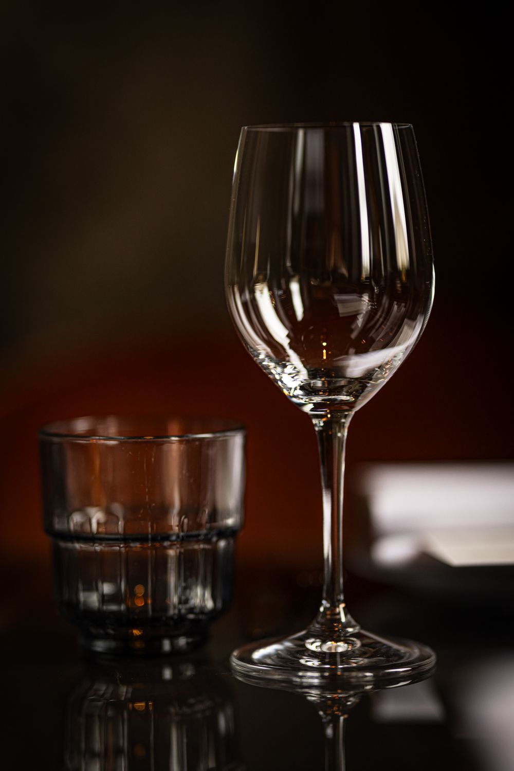 Empty wine glass and low tumbler on a dark bar table, softly lit against a blurred background