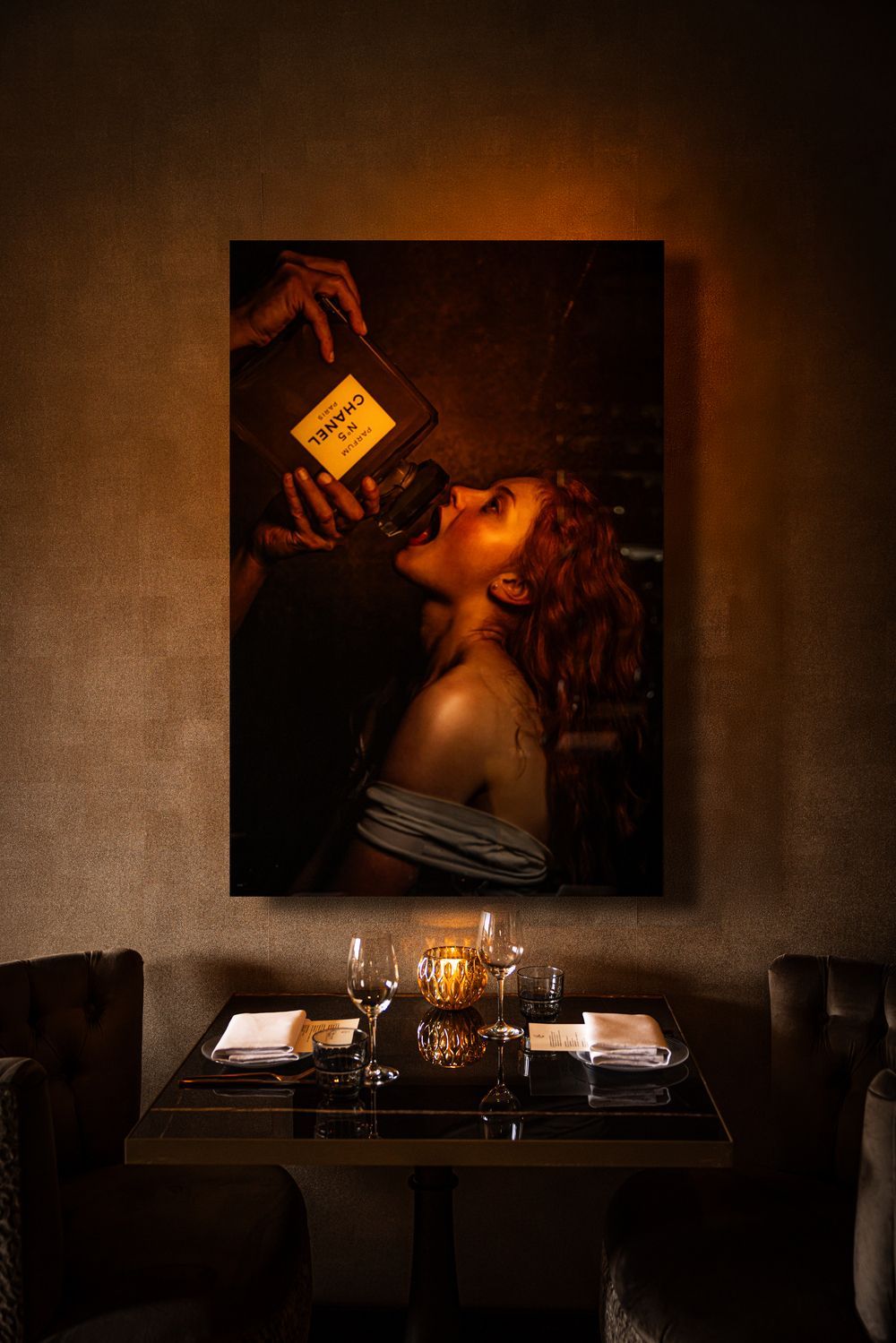 Woman in warm candlelit restaurant gazes up at a wine bottle poster above a set dining table.