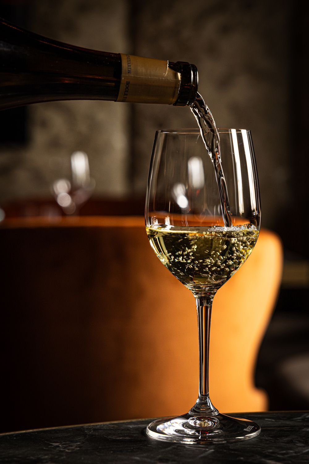 Wine being poured into a clear wine glass on a dark table, with warm ambient lighting.