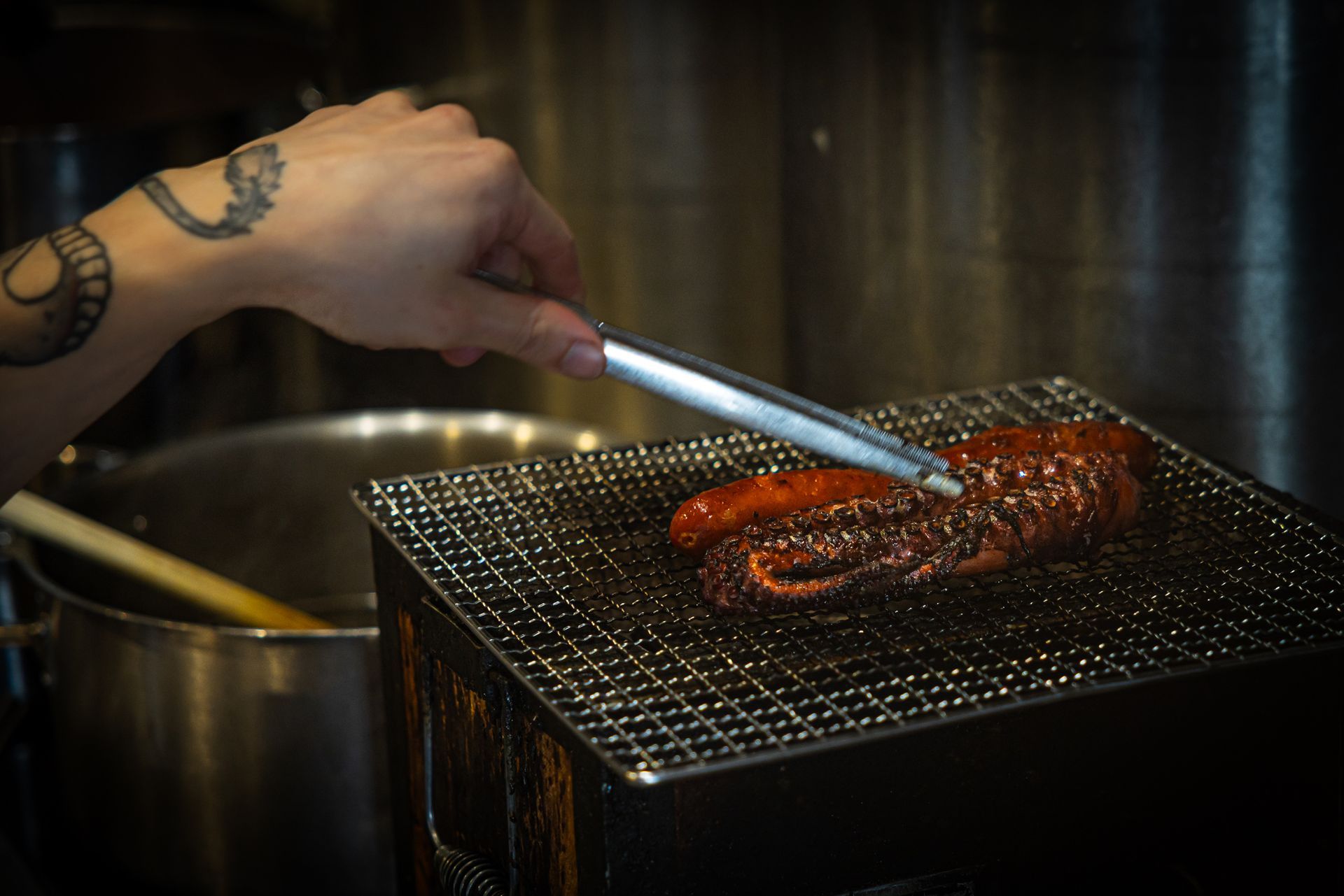 Hand tattooed hand uses tongs to grill reddish meat over a wire rack