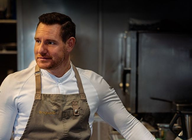 Chef in a beige apron and white shirt standing in a professional kitchen, looking to the side.