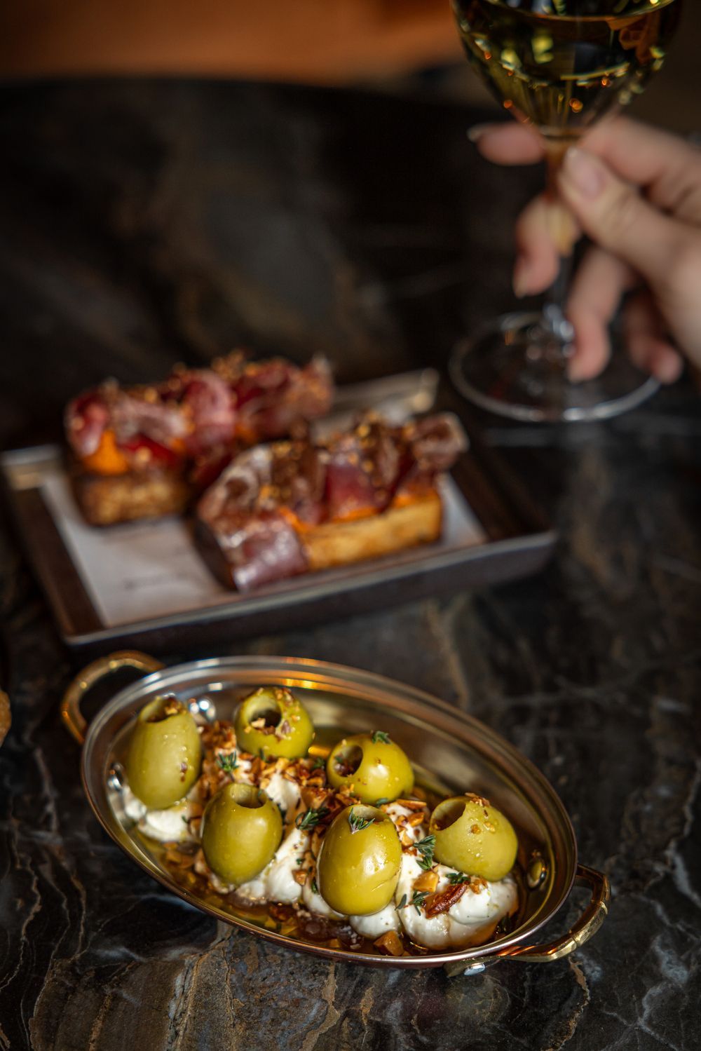 Plate of glazed meat bites and a pan of potatoes with olives, beside a hand holding a wine glass