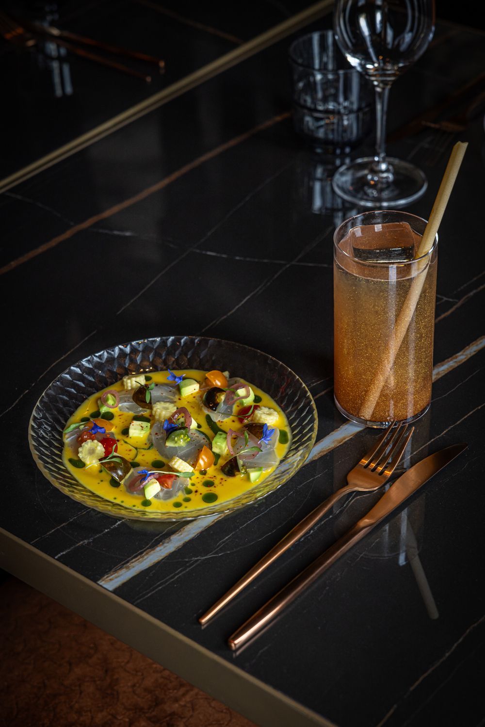 Colorful appetizer in a small skillet beside a tall cocktail on a dark table with chopsticks.