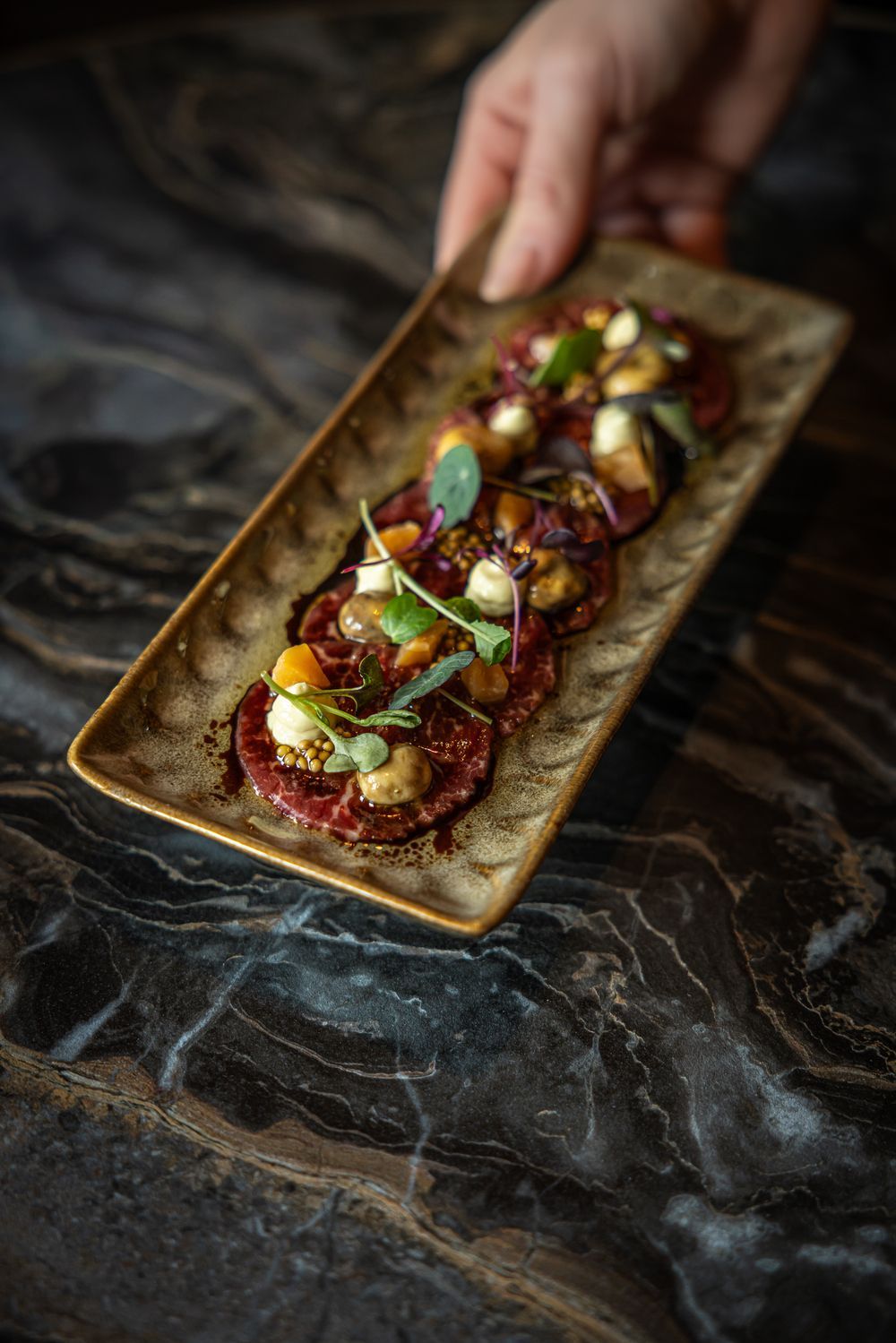 Hand placing a rectangular platter of garnished bite-sized appetizers on a dark marble surface