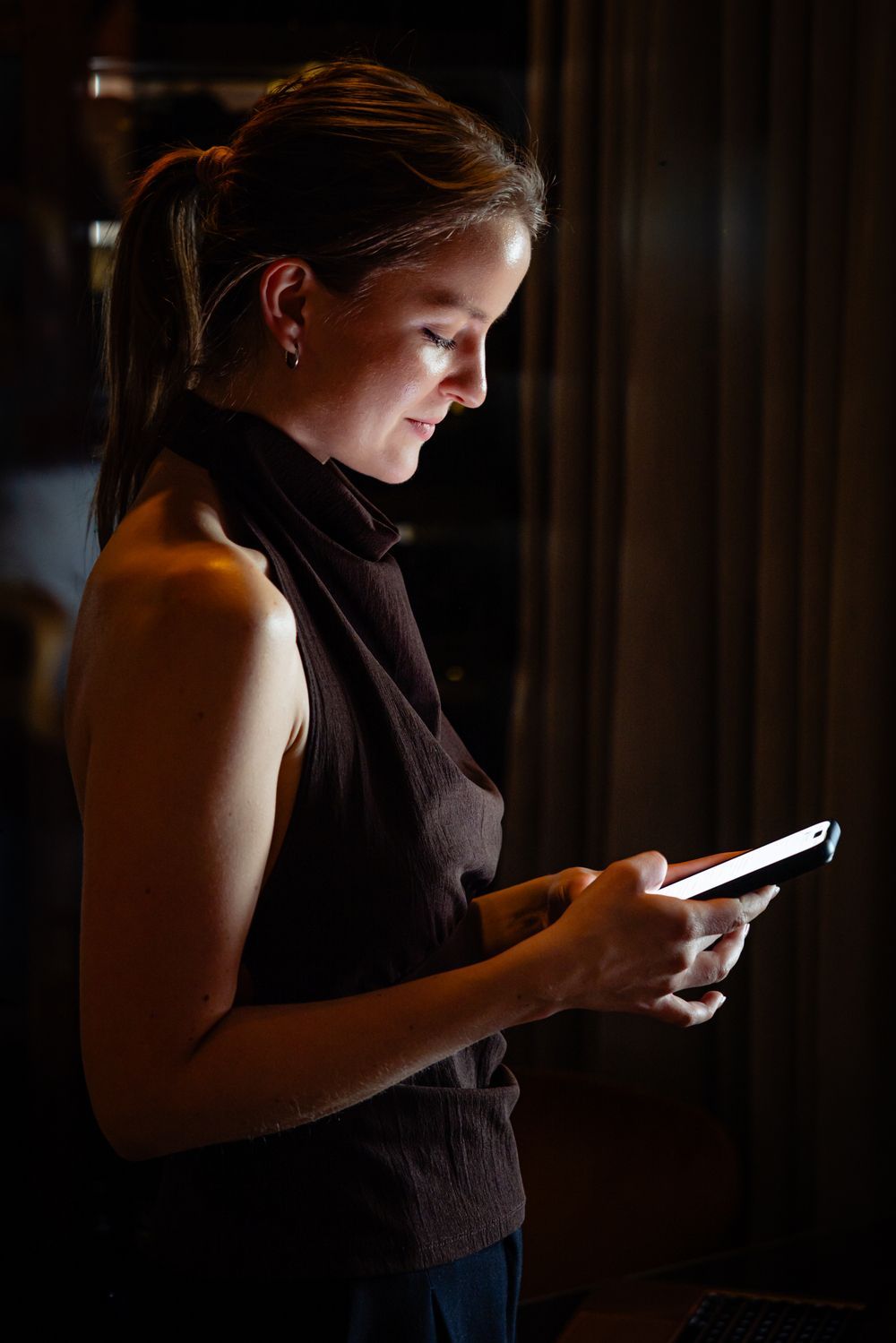 Person in a dark room looking at a glowing smartphone screen by the window
