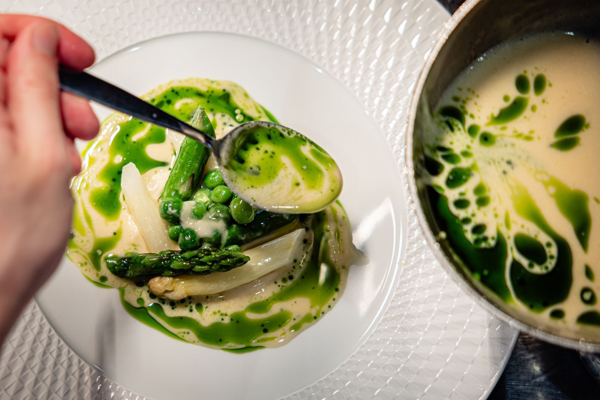 Hand stirring green-sauced vegetable risotto in a white bowl beside a pot of creamy soup