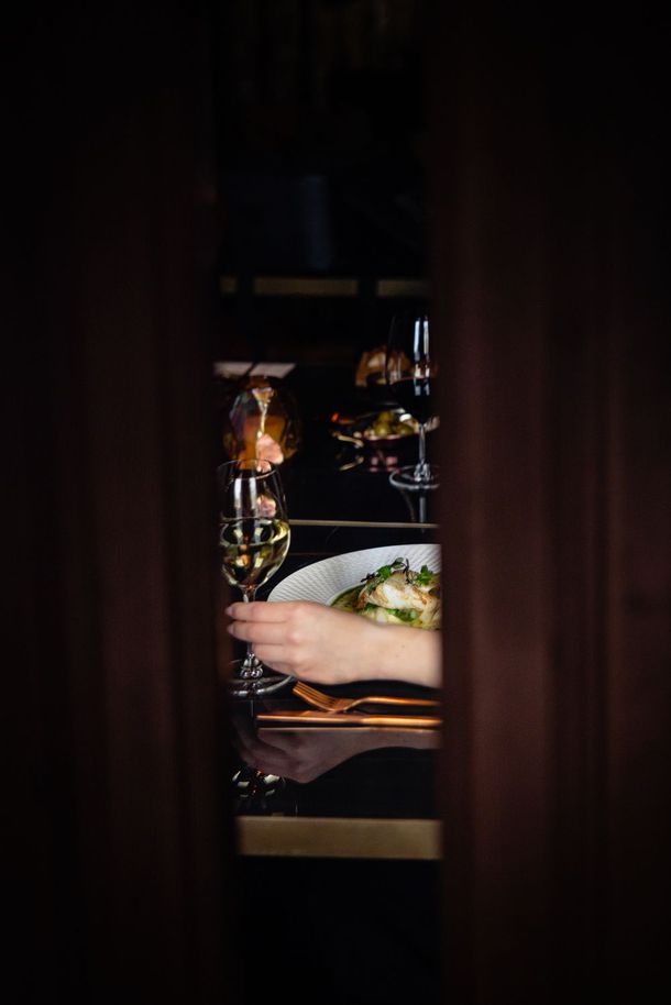Hands lifting a drink at a dimly lit restaurant table, seen through a narrow gap in dark curtains