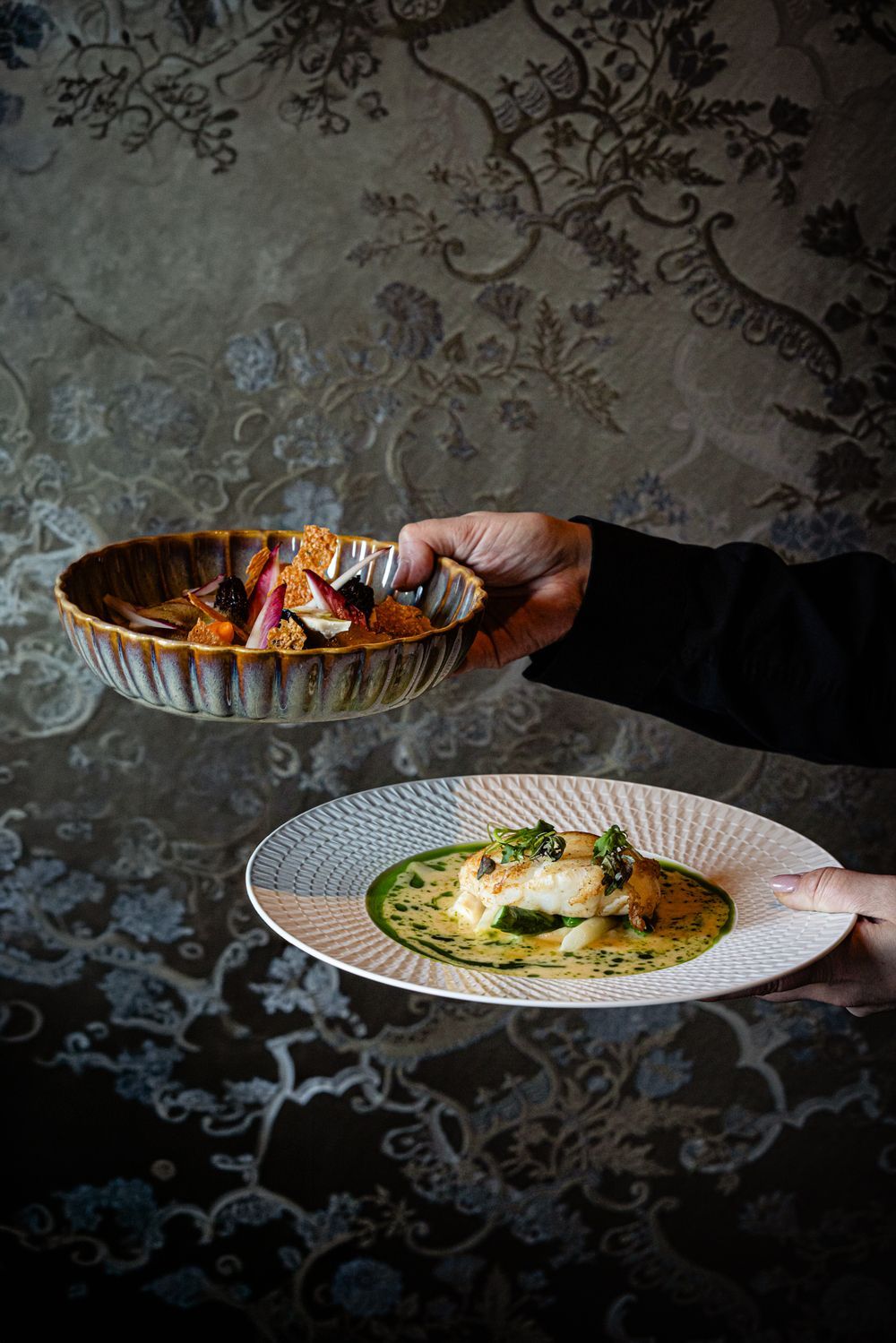 Hands serving food onto a white plate in a dimly lit restaurant setting
