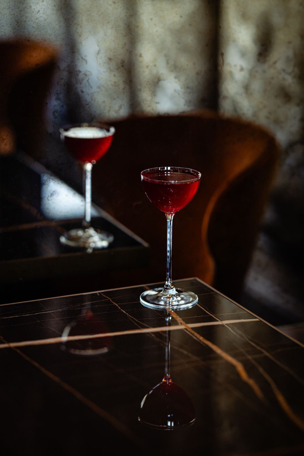 Two red cocktails on a glossy table beside a blurred chair and curtain.