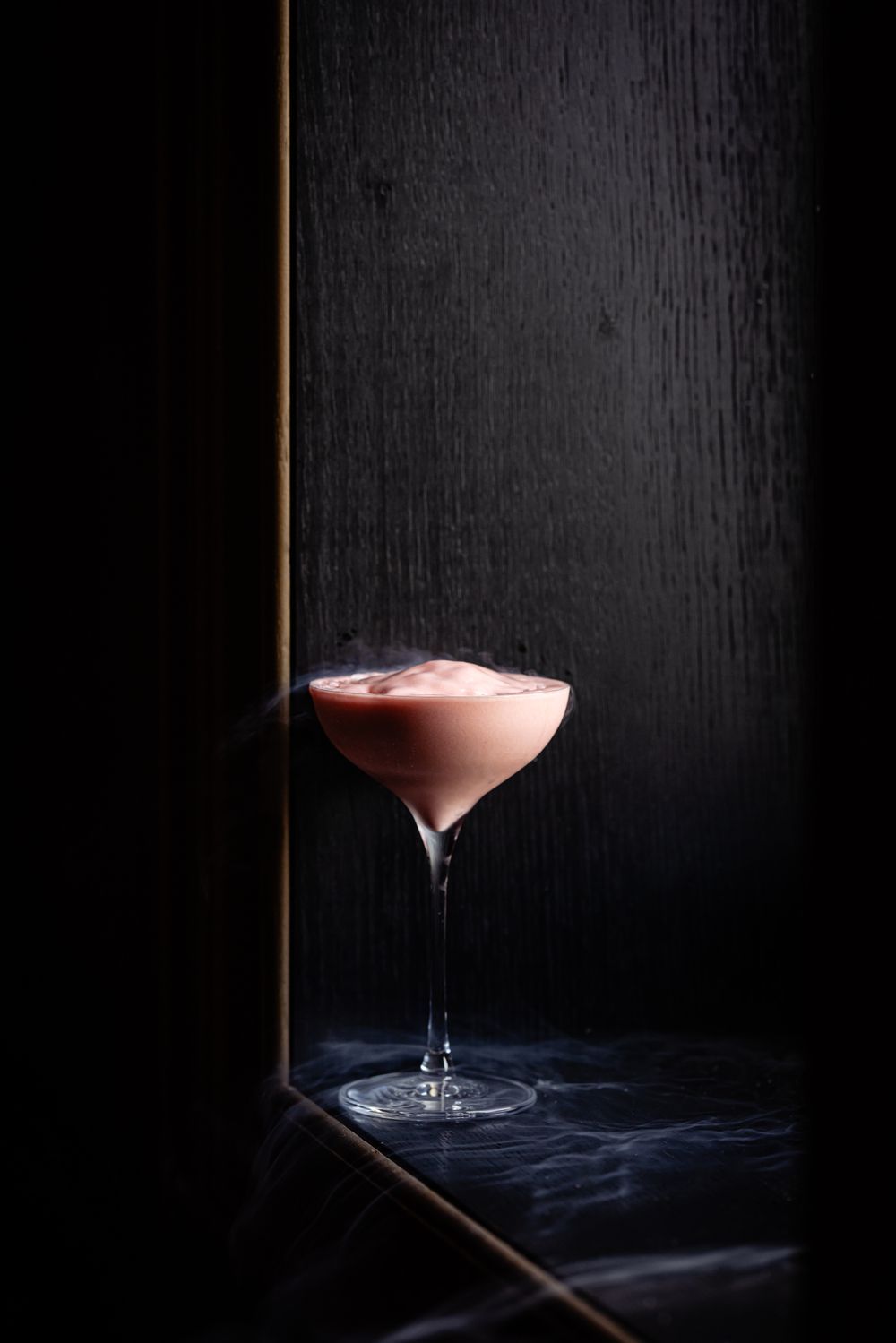 Pink cocktail in a stemmed glass on a dark surface, lit against a black background.