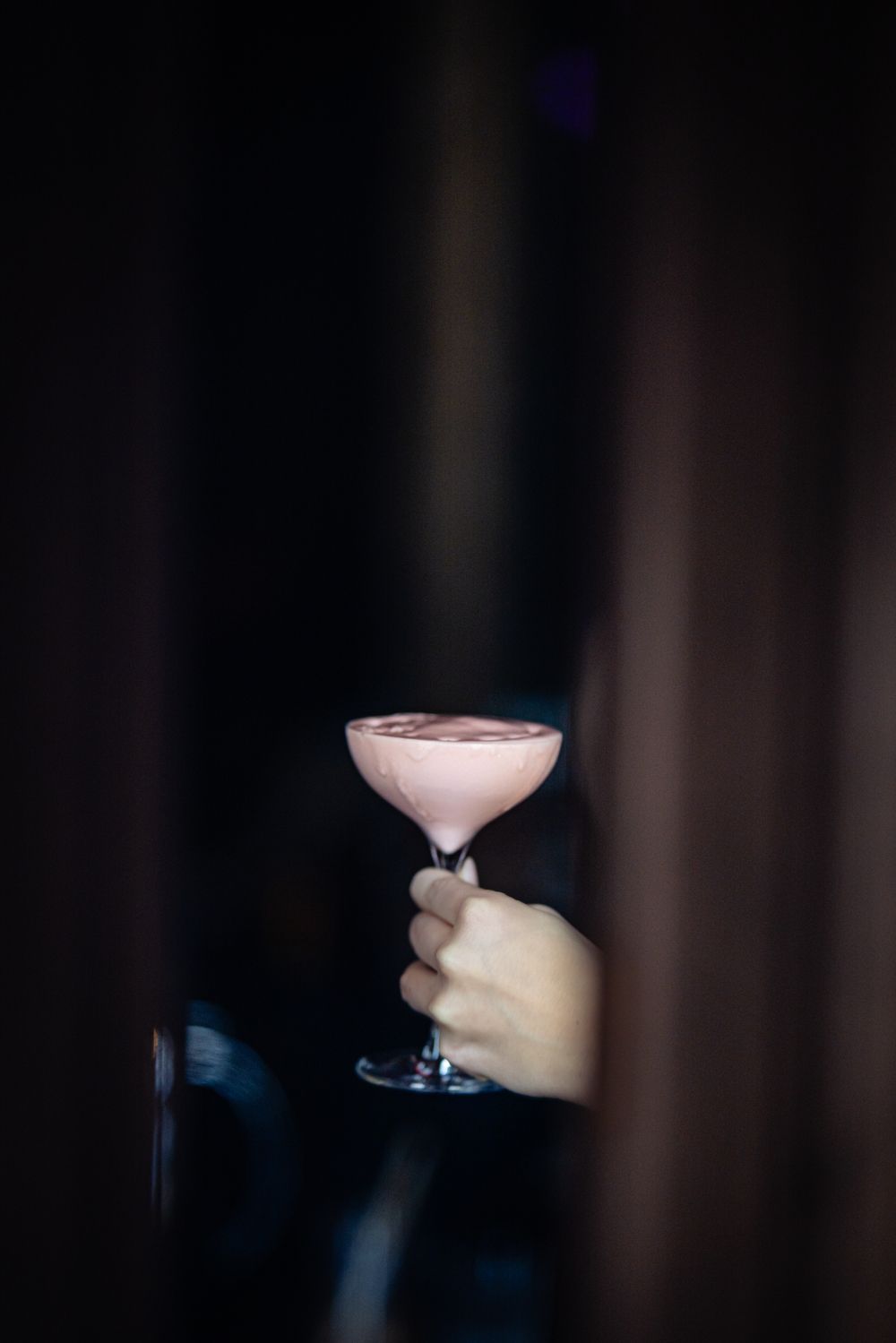Hand holding a pink cocktail in a coupe glass against a dark, blurred background