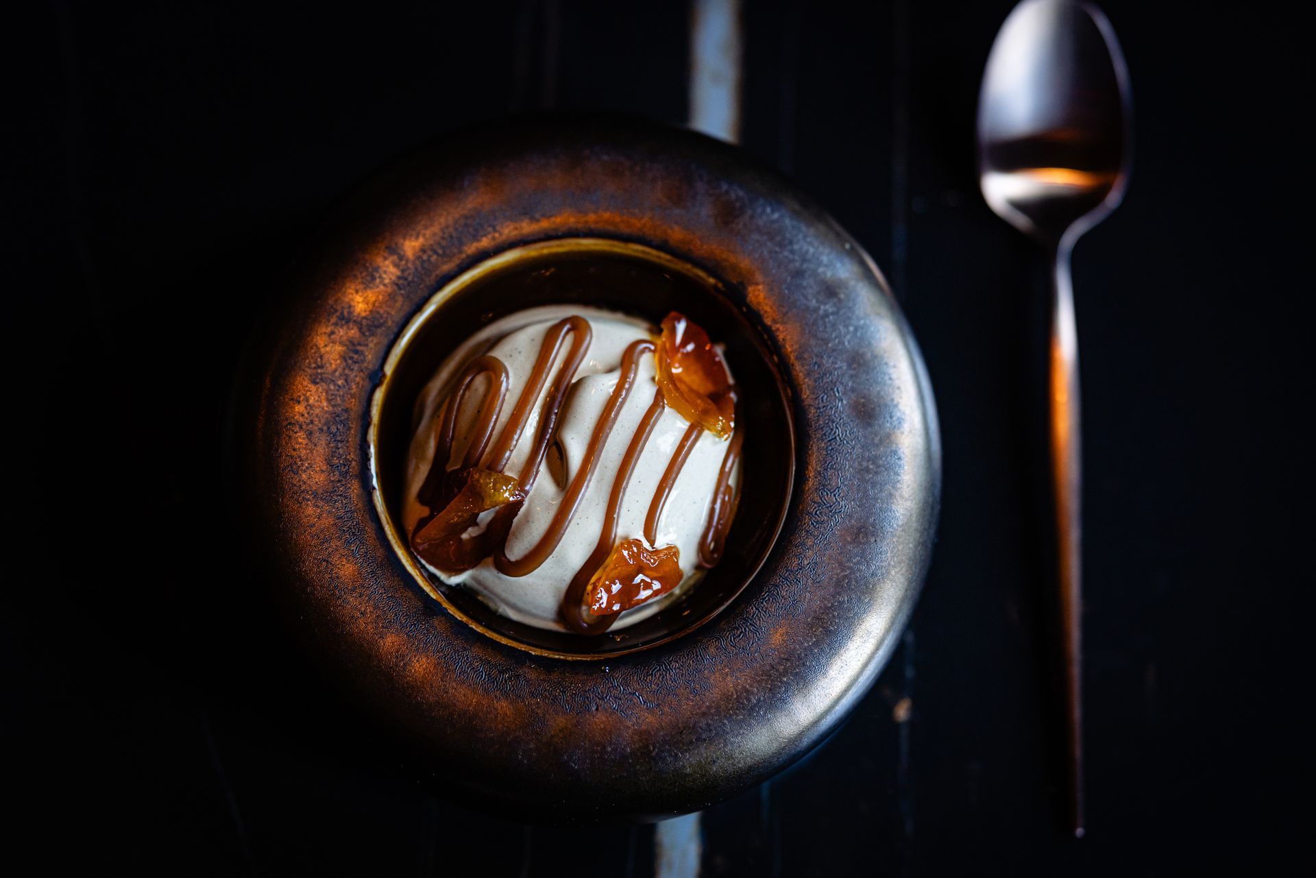 Top-down view of a dessert in a dark cup with caramel drizzle, beside a spoon on a black background