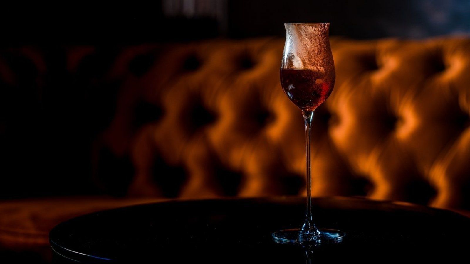 A single wine glass on a dark table before a warmly lit tufted lounge backdrop.