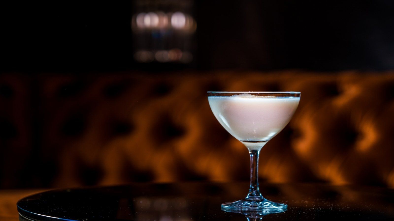 White cocktail in a martini glass on a dark table against a warm, blurred lounge backdrop