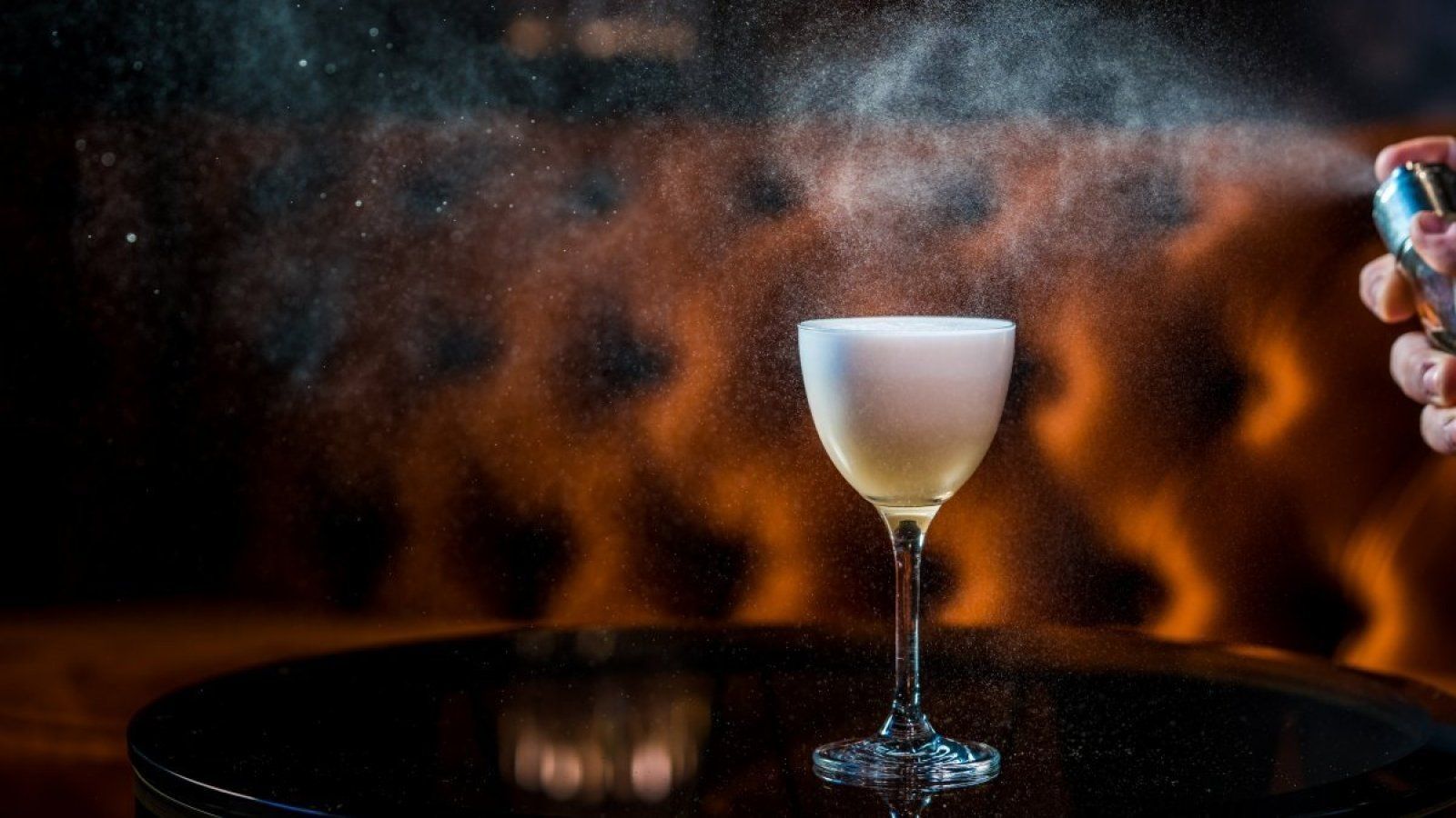 Cocktail glass on a tray with orange flames in the background, with a hand holding a torch at right