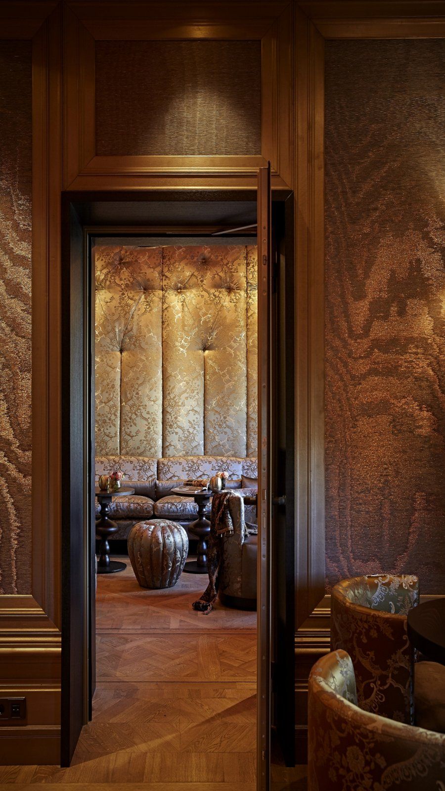 Ornate wood-paneled hallway opening into a warm-lit room with patterned walls and upholstered chairs