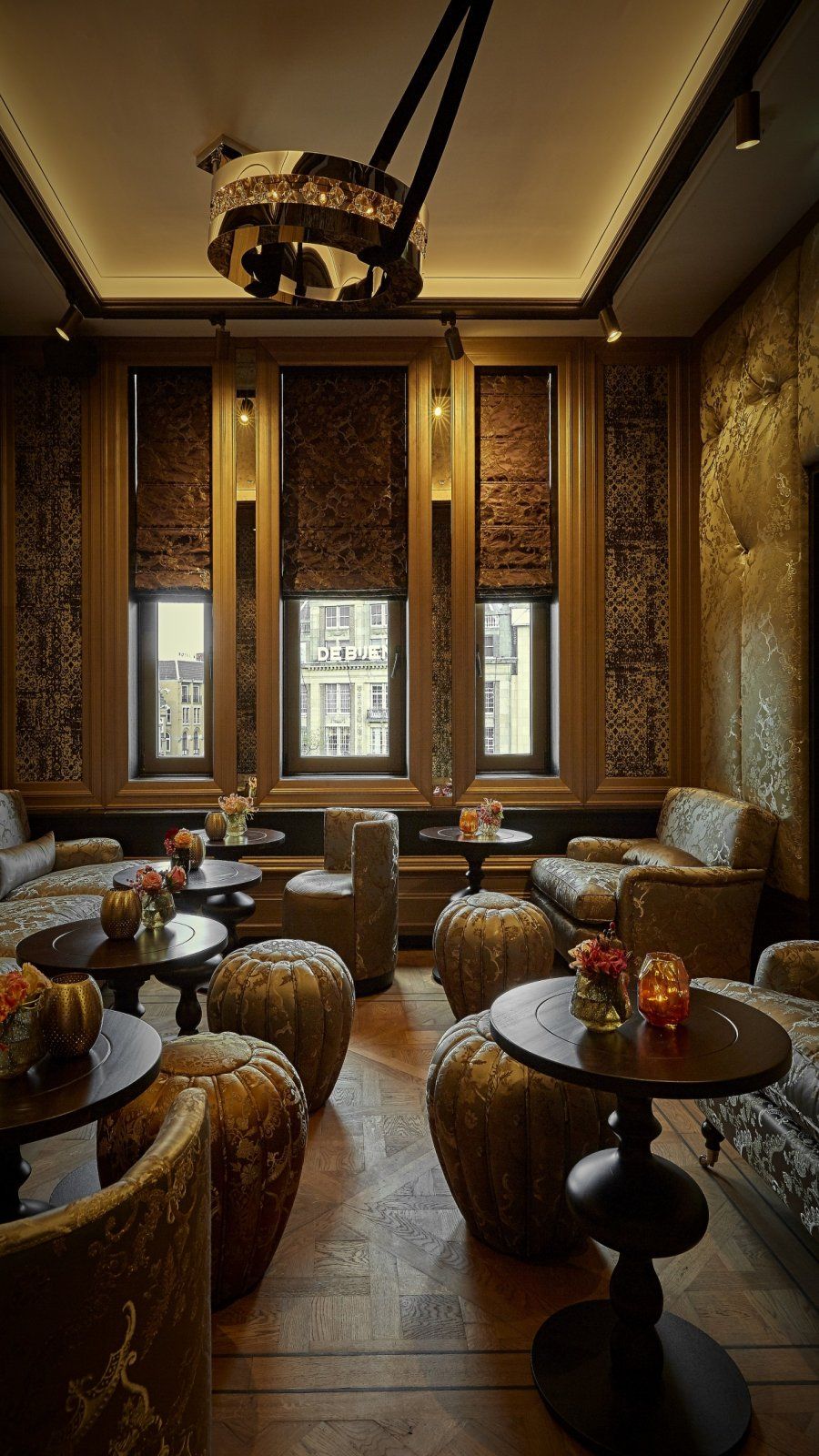 Elegant lounge with ornate gold decor, round tables, patterned chairs, and tall windows with city view.