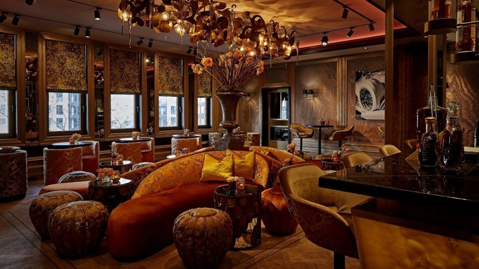Elegant lounge with warm orange seating, chandeliers, and large windows in a dimly lit room