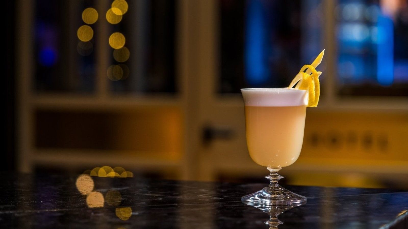 Cocktail in a stemmed glass with pineapple garnish on a bar counter, warm bokeh lights in the background