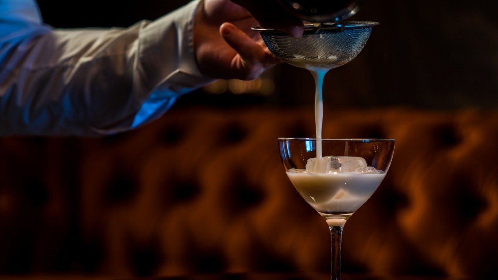 Hand pouring a blue cocktail into a martini glass in a dimly lit bar