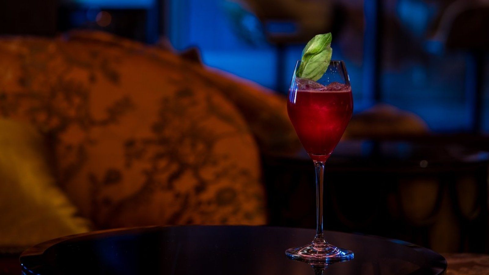 Red cocktail in a stemmed glass on a table with a lime garnish, against a blurred lounge backdrop