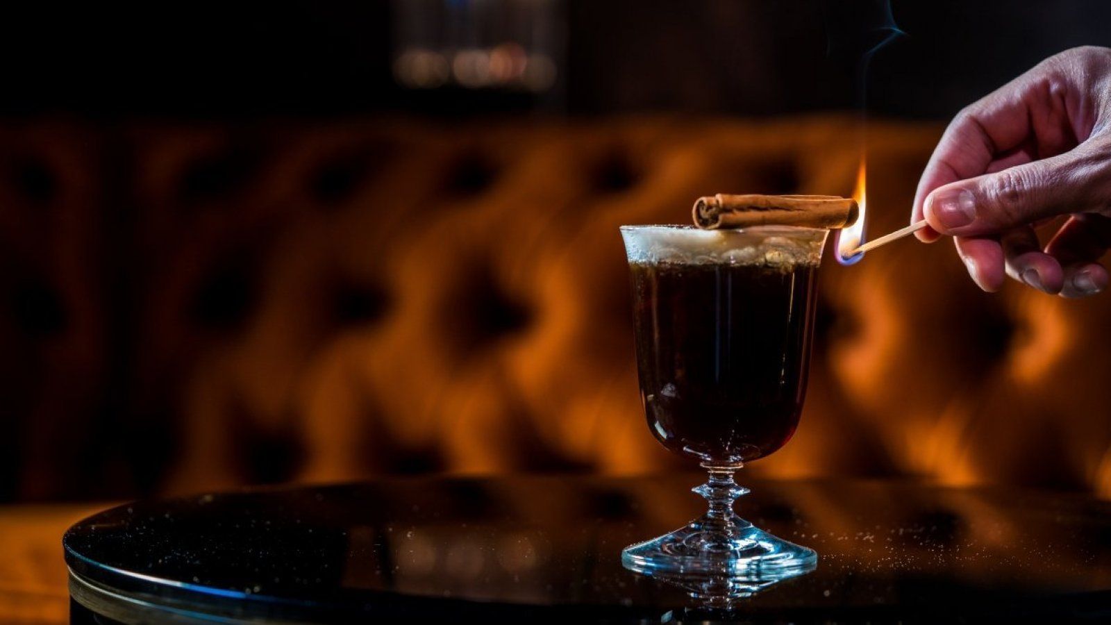 Hand placing a lit match near a glass of dark drink on a tray in a dimly lit bar