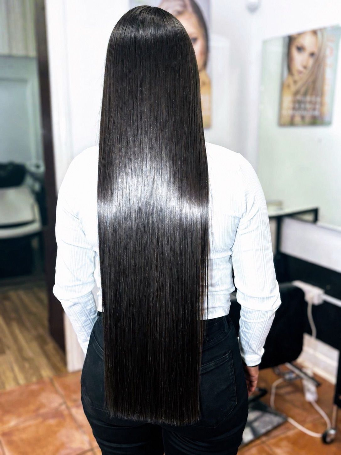 Persona con cabello largo, liso y negro de pie en un salón de belleza o estudio de peluquería, de espaldas a la cámara.